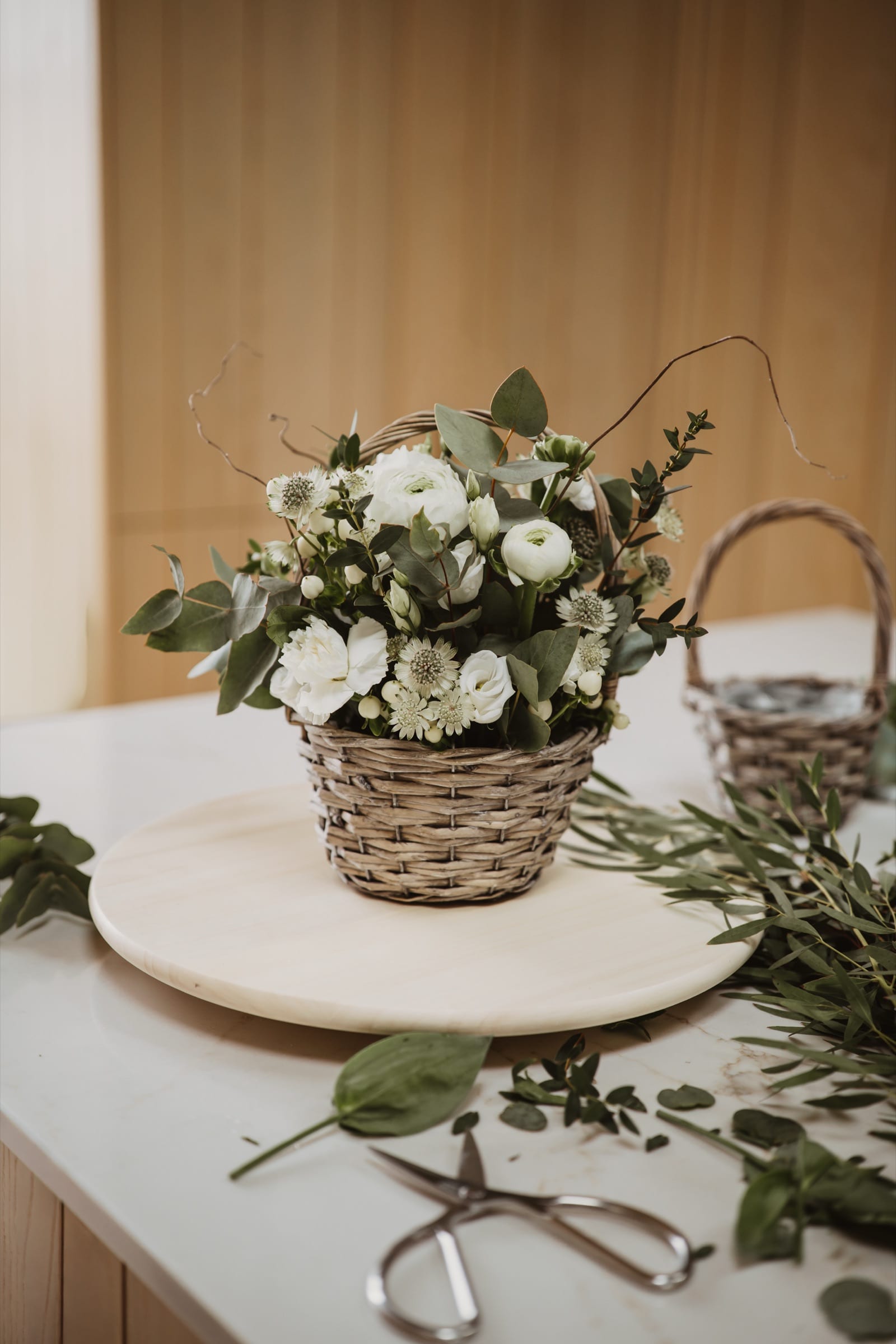 A basket of white ranunculus and eucalyptus on a work surface, with scissors and cut foliage laid around it.