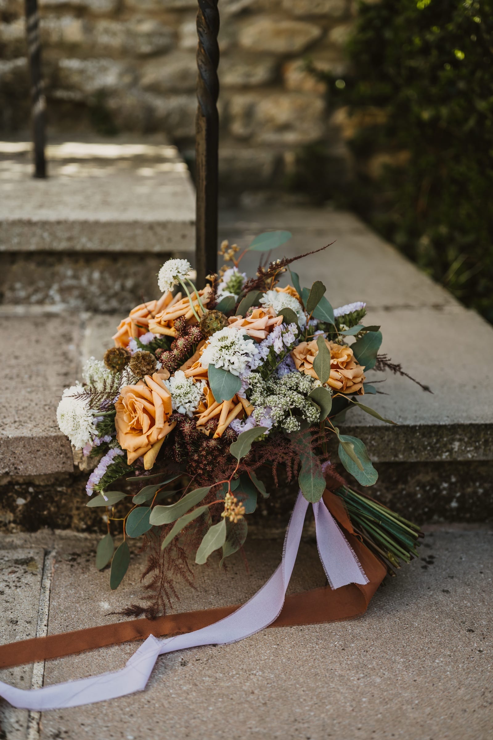 A wedding bouquet of apricot roses and dried grasses resting on stone steps, ribbons trailing.