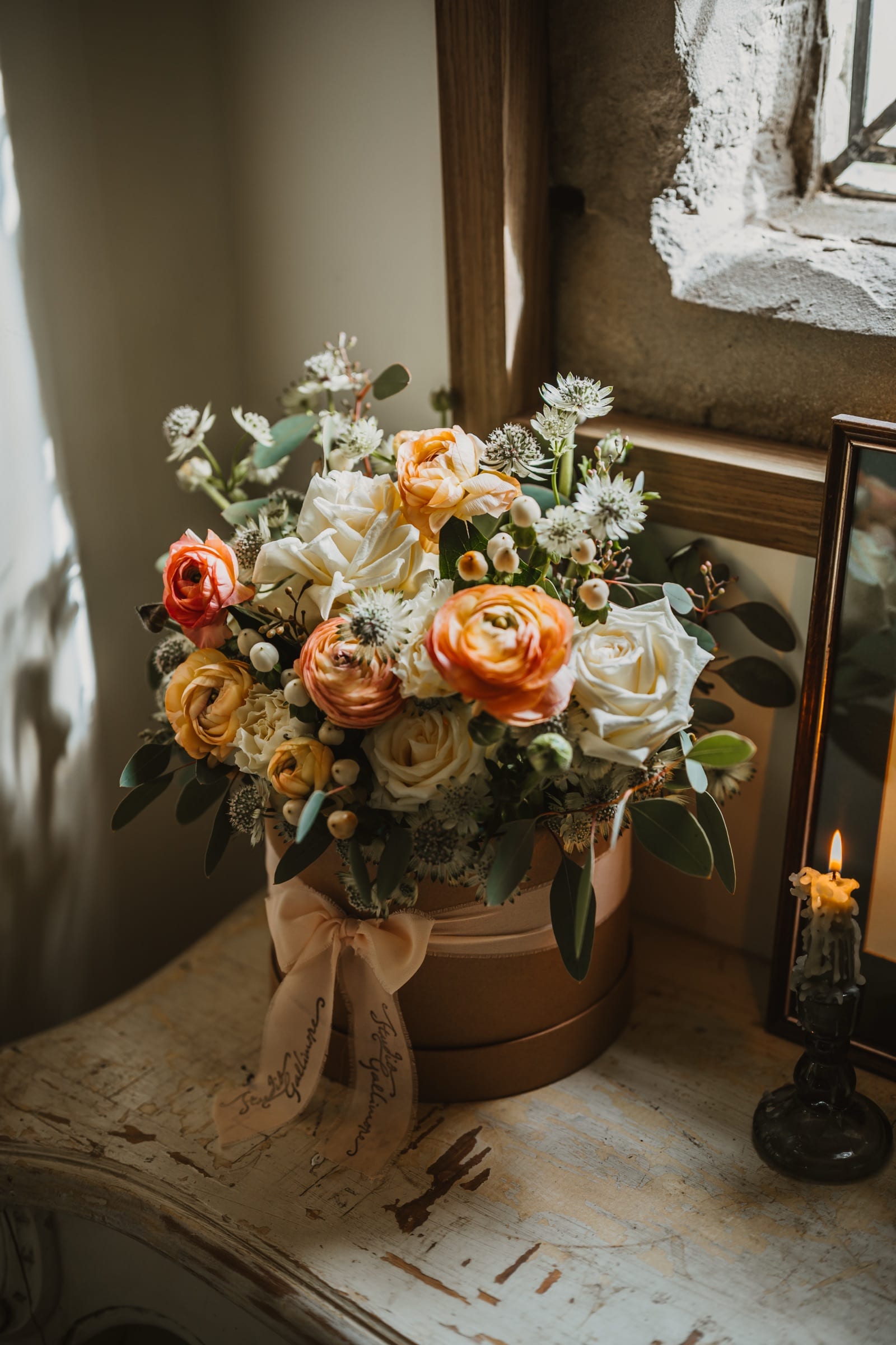 Close view of a hatbox arrangement of ranunculus and roses by a stone window, candle burning to one side.