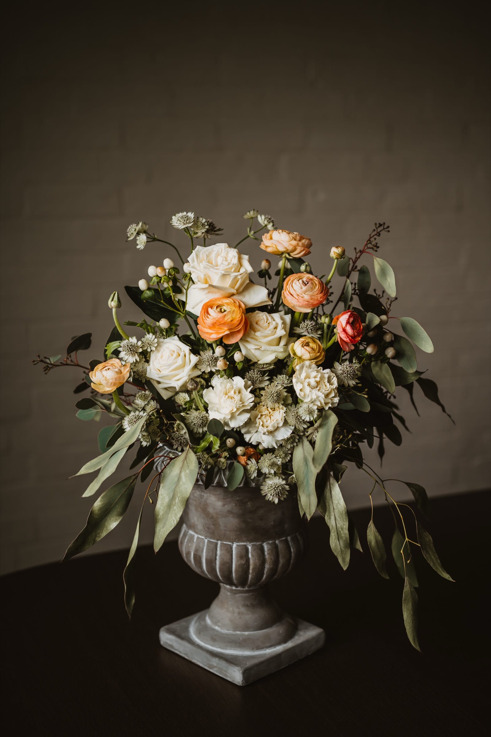 An arrangement of peach ranunculus, white roses and eucalyptus in a fluted stone urn against a dark brick wall.