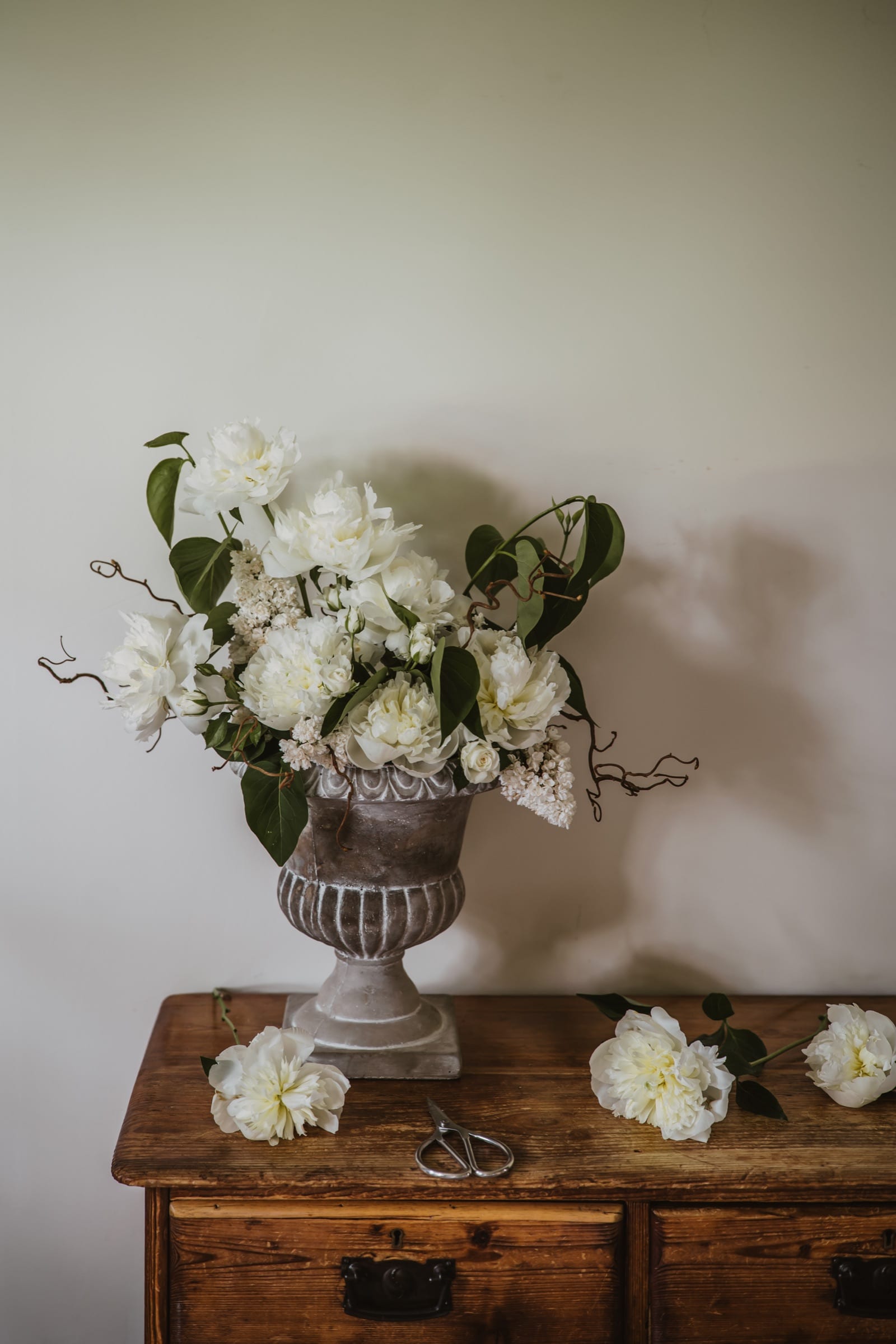White peonies in a stone urn on an antique chest of drawers, loose blooms and scissors laid beside it.