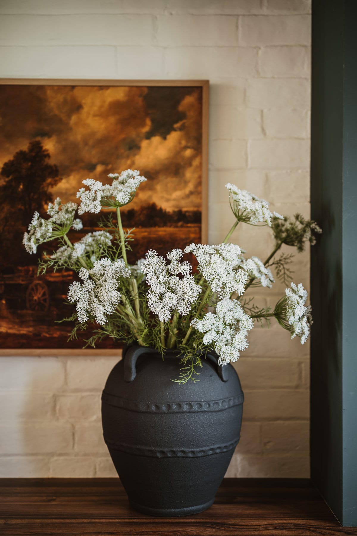 Wild carrot flowers in a black textured urn on a wooden surface, beneath a landscape painting on a painted brick wall.