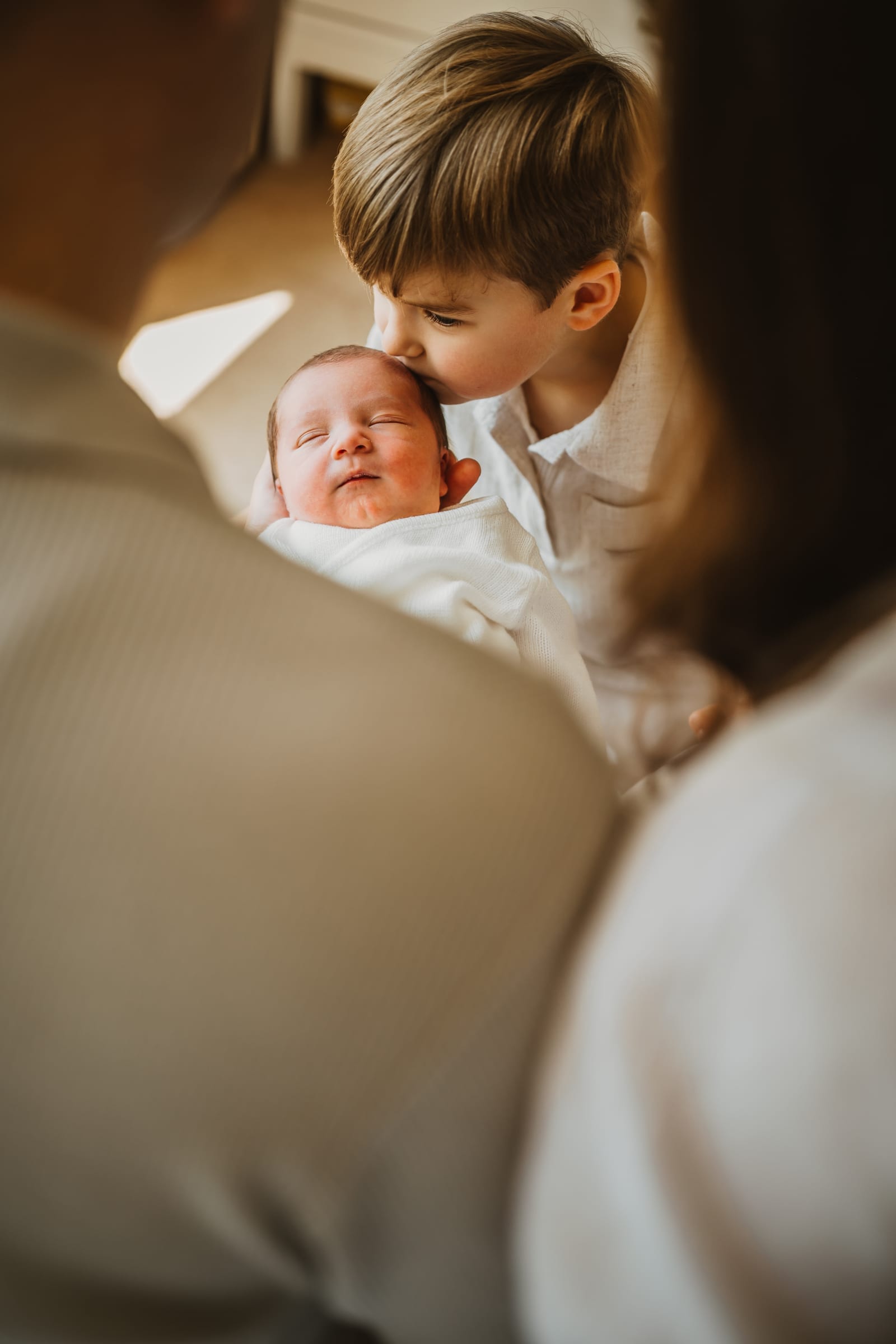 A small boy kisses his newborn sibling's head, the baby wrapped in white and held between two parents seen from behind.