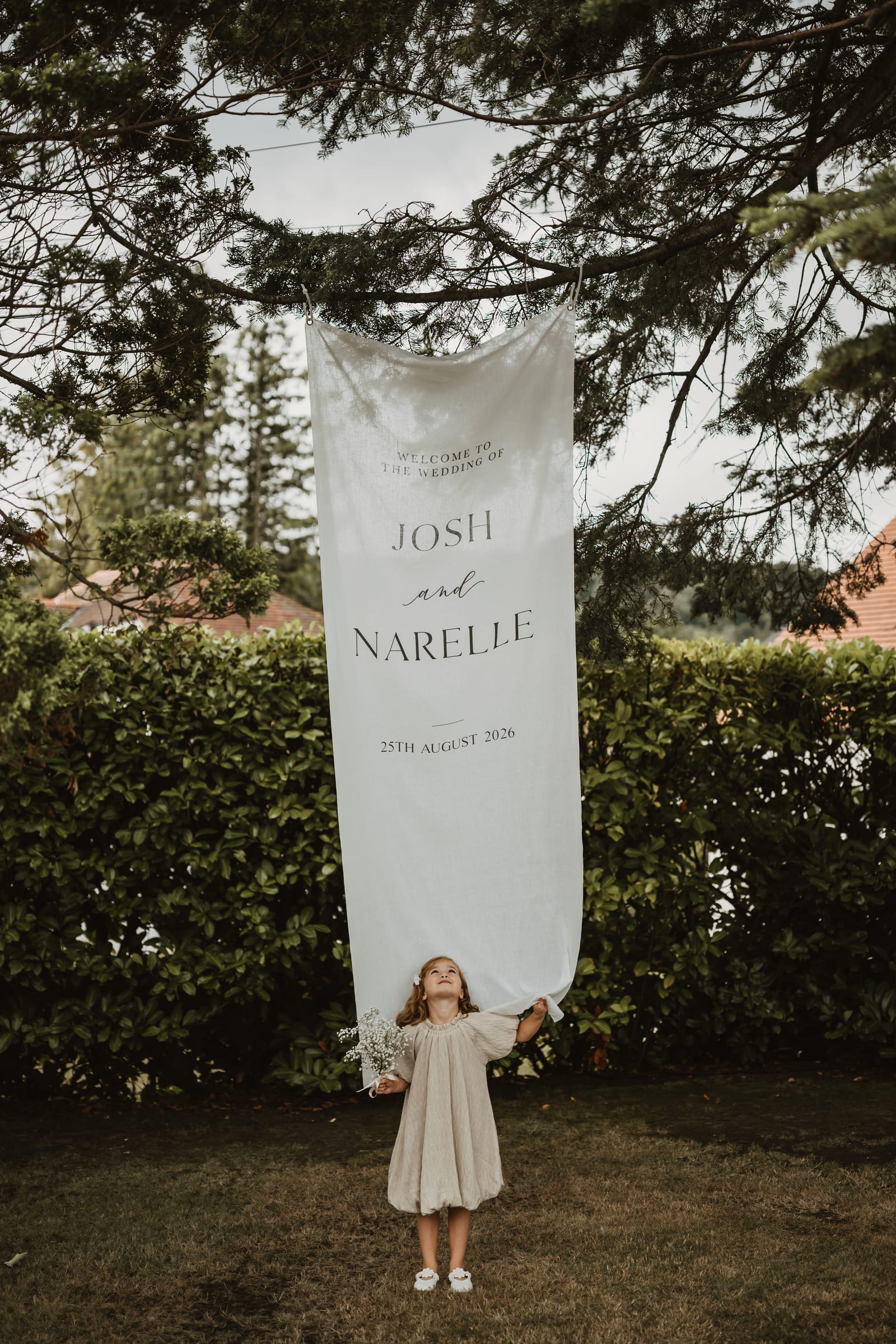 A flower girl holding a posy looks up at a tall linen welcome banner hung from a cedar branch over a hedge.