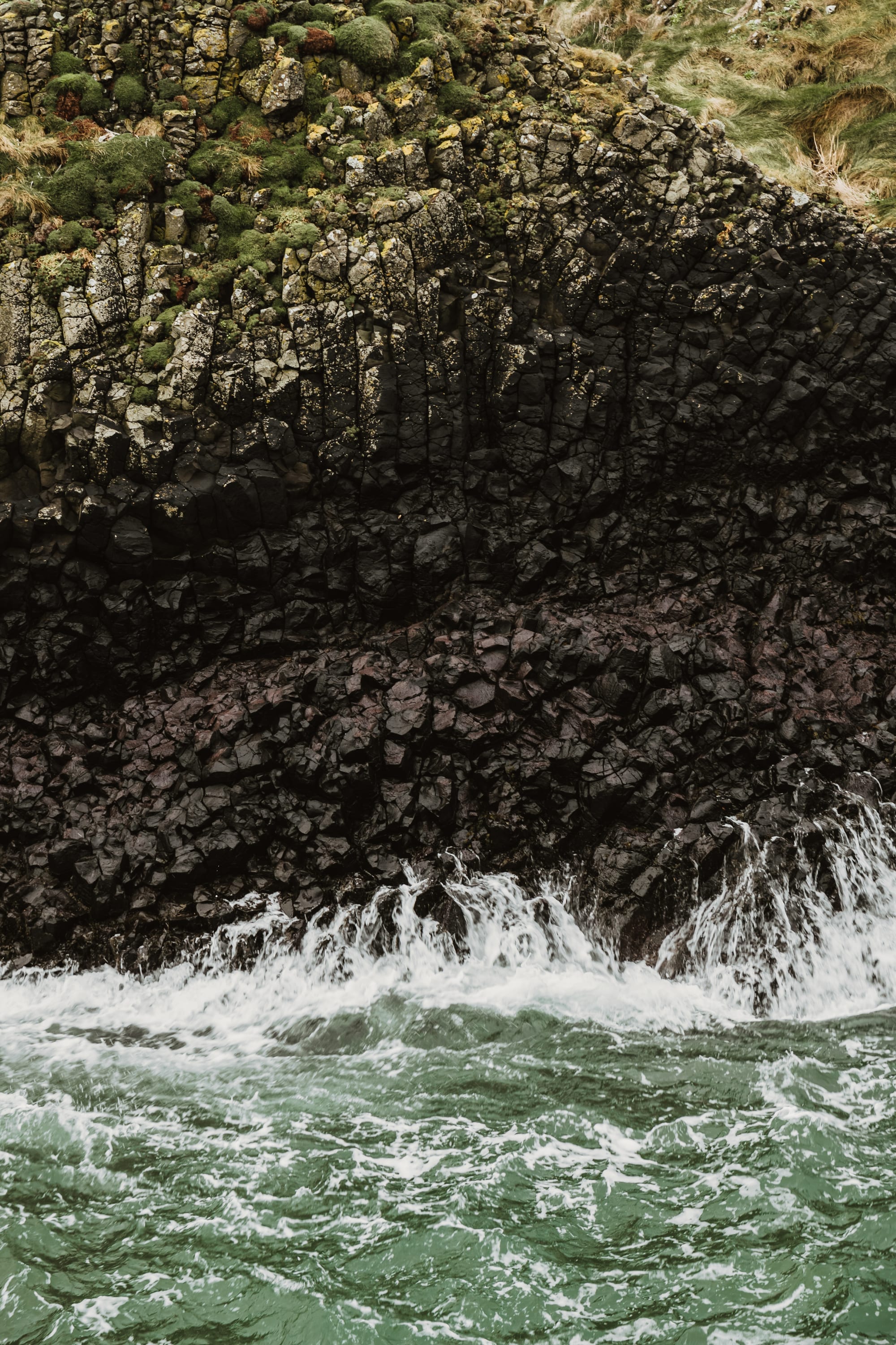 Moss and grass on a wall of fractured basalt columns, with white water breaking into green sea at its foot.