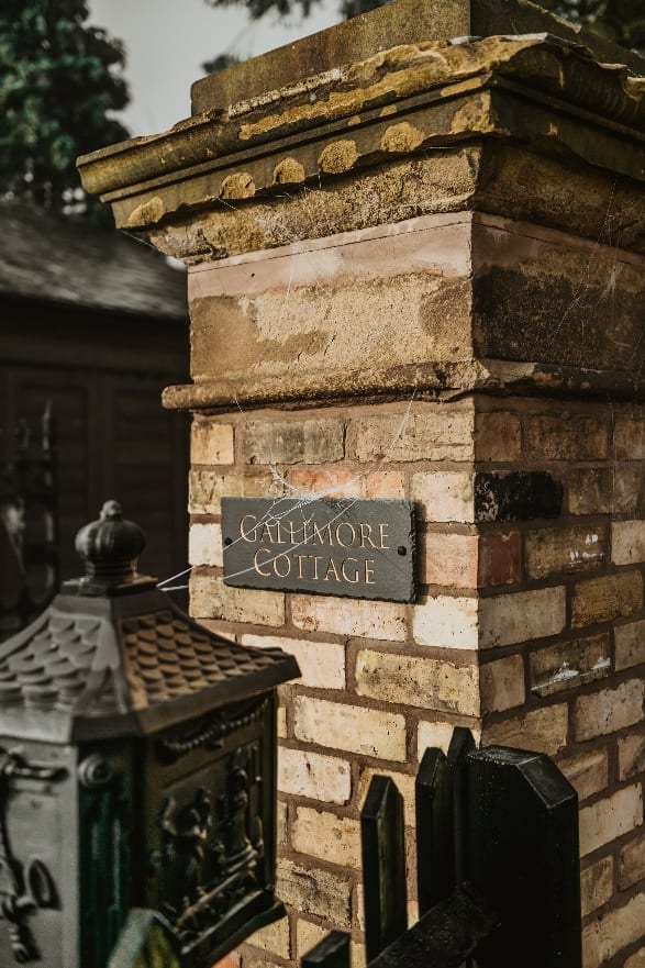 A brick and stone gate pier carrying a slate sign reading Gallimore Cottage, an ornate dark green cast iron box in the foreground.