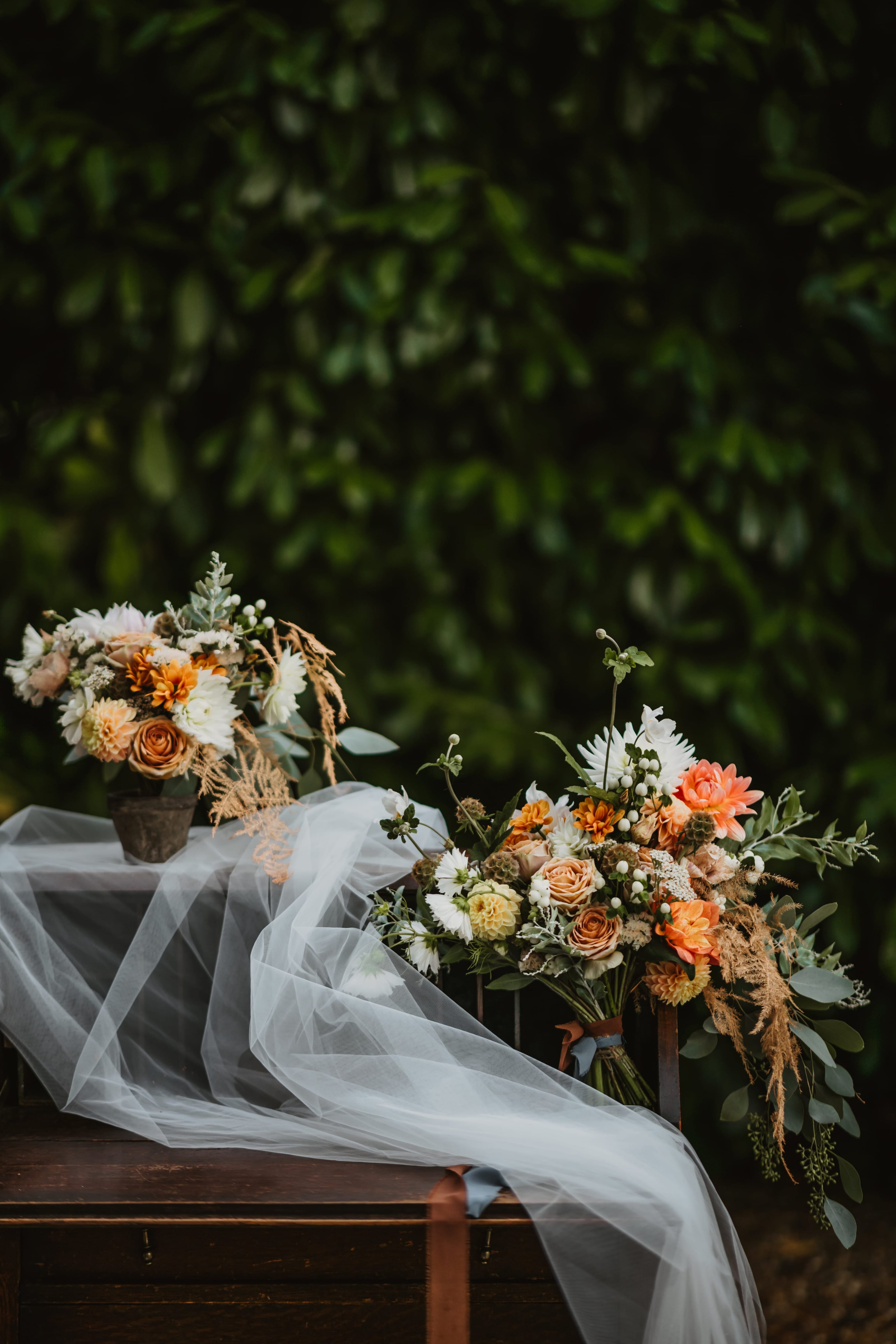 A bridal bouquet of peach roses, white blooms and trailing foliage resting on a bench, its veil drawn out beside it against dark greenery.