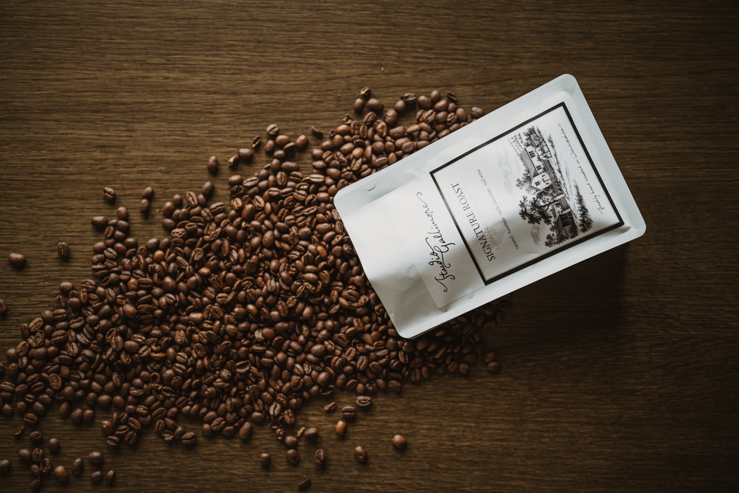 Seen from above: a bag of Studio Gallimore Signature Roast lying on a dark wooden table in a wide spill of roasted beans.