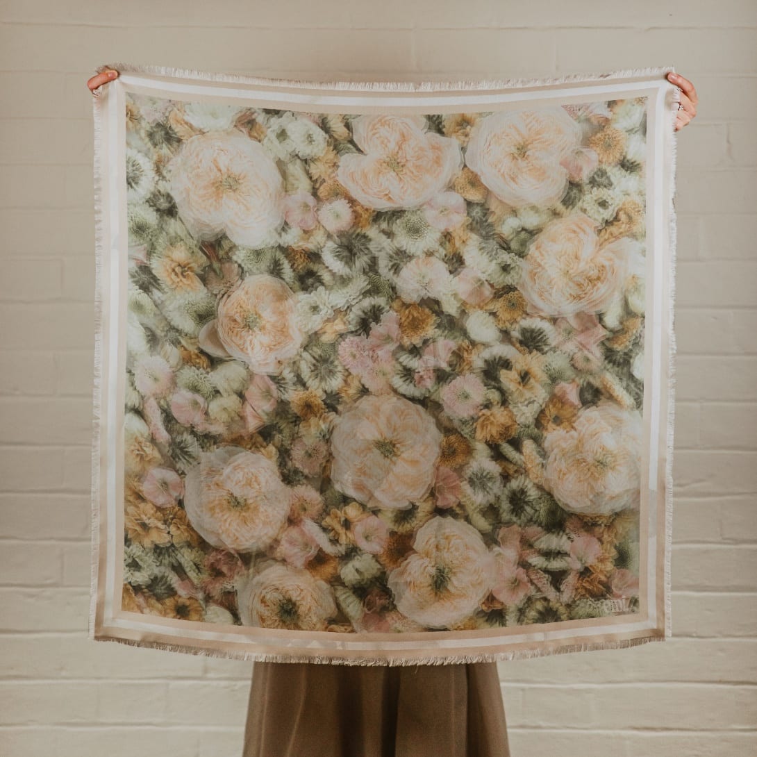 Bailie, a silk scarf held against a painted brick wall, filled edge to edge with peach, blush and ivory roses and finished with a pale frayed edge.