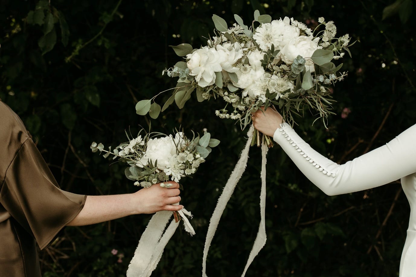 Two hands exchanging bouquets of white peonies, scabiosa and eucalyptus, one tied with long ivory ribbons, against dark foliage.