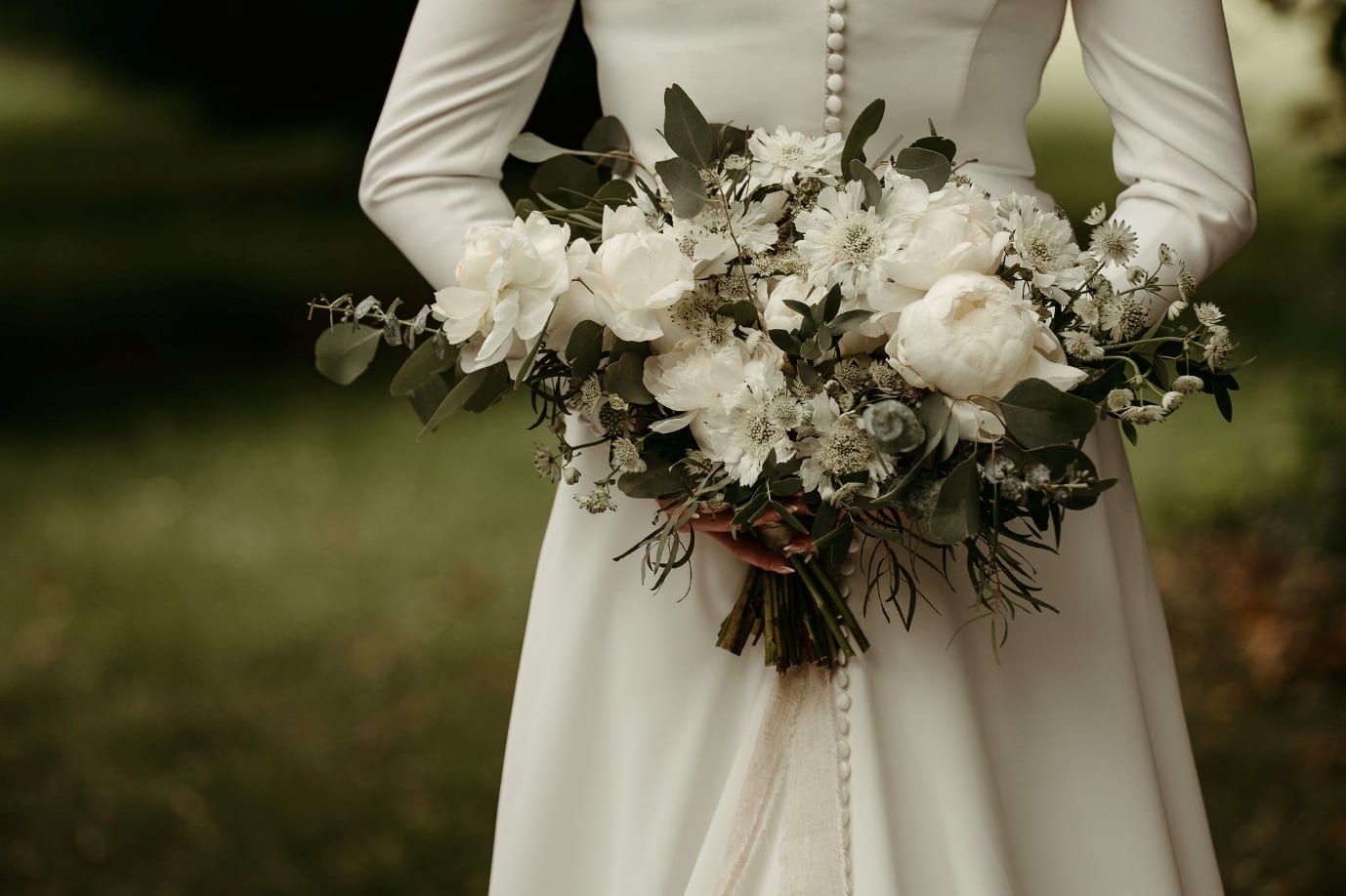 A bride in a long-sleeved ivory dress with covered buttons, holding a green and white bouquet of peonies and eucalyptus.