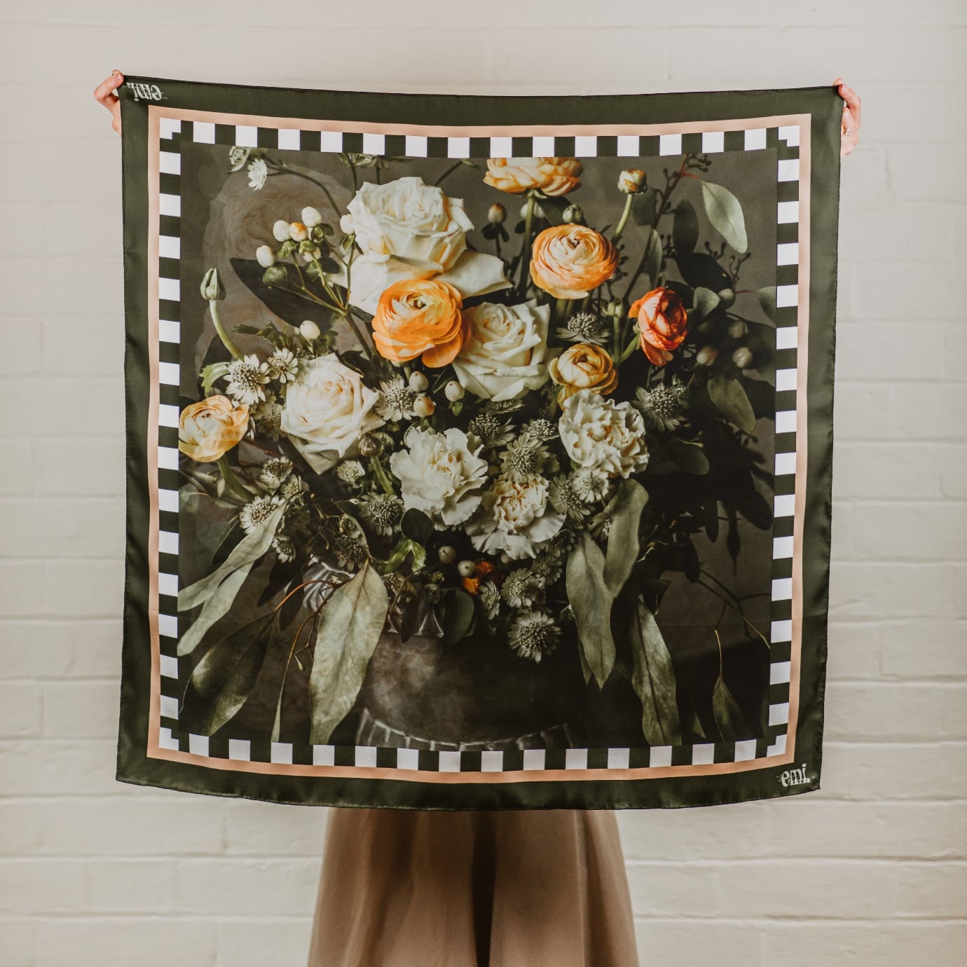 Bryn, a silk scarf printed with cream roses and orange ranunculus in a dark urn, framed in olive with a black and white checkerboard border.