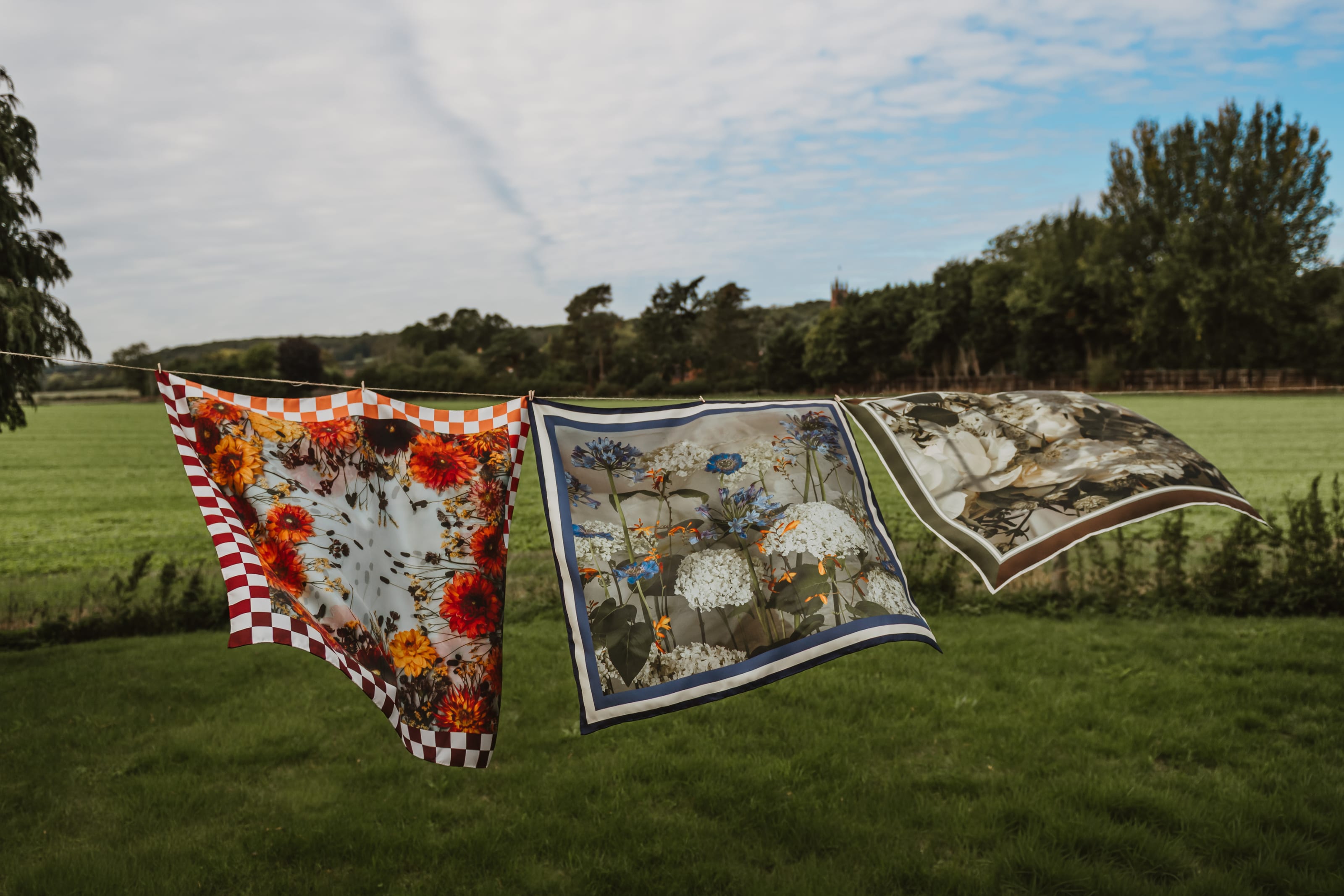 Three Emi silk scarves pegged to a line across a field under a pale sky: dahlias in a red check border, hydrangea and agapanthus in navy, white peonies in brown.