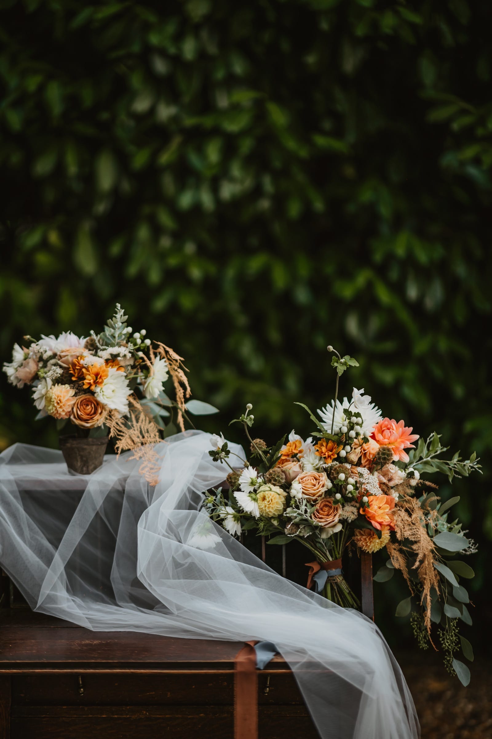 A bridal bouquet of peach roses, white blooms and trailing foliage resting on a bench, its veil drawn out beside it against dark greenery.