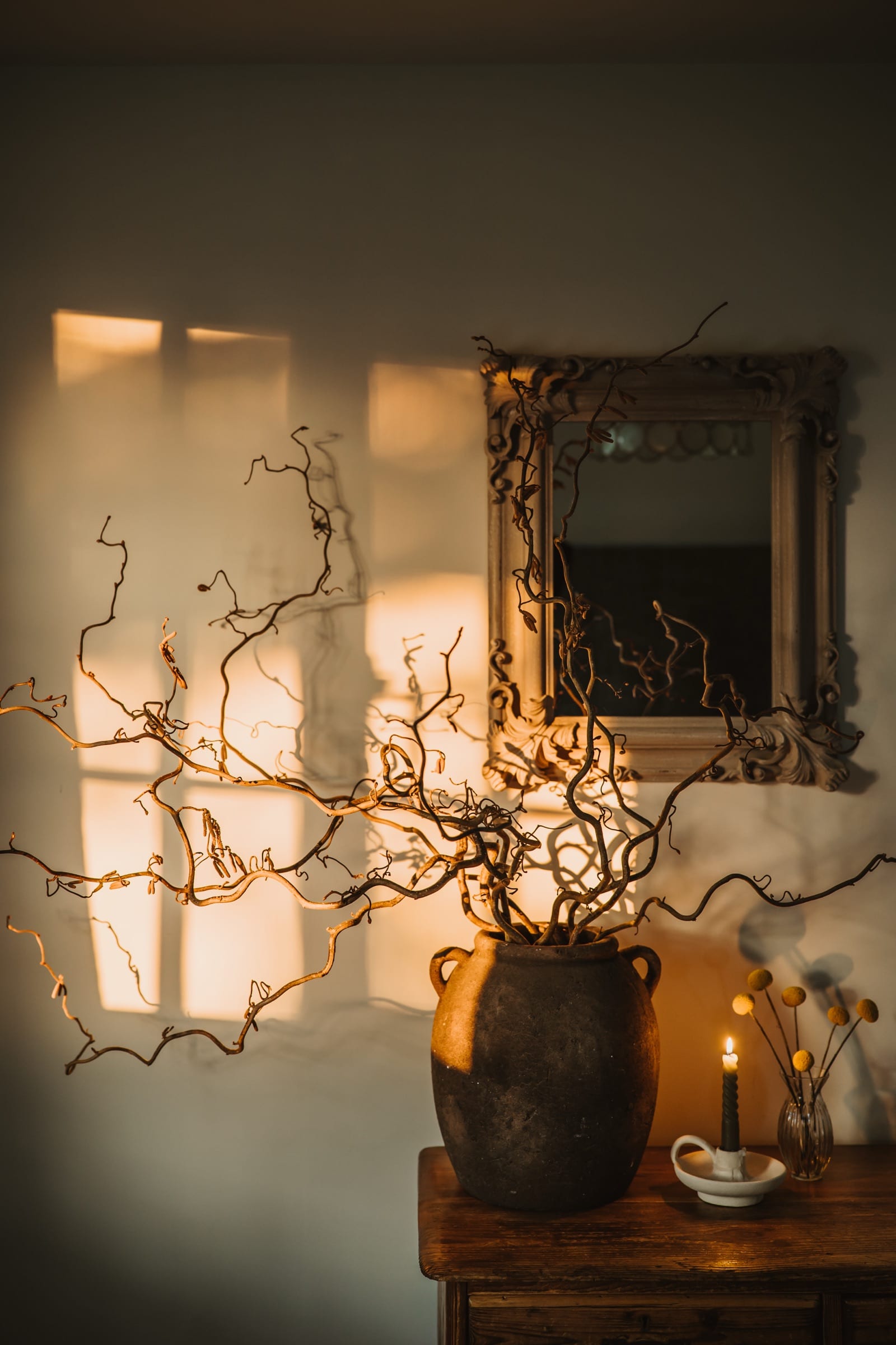 Contorted hazel branches in a stoneware urn on a wooden sideboard, beneath a gilt mirror, in low afternoon light.