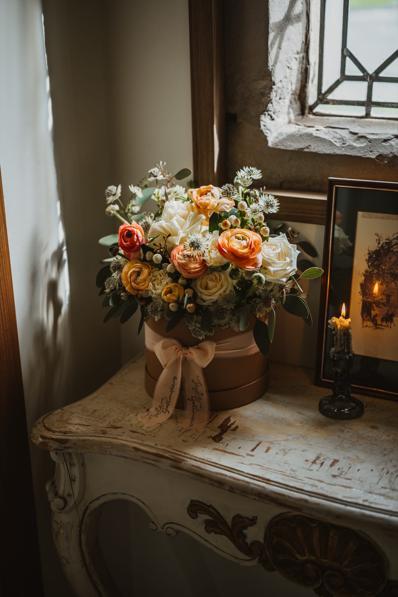 A hatbox arrangement of peach and cream flowers on a painted console beside a leaded window, with a lit candle and a framed picture.