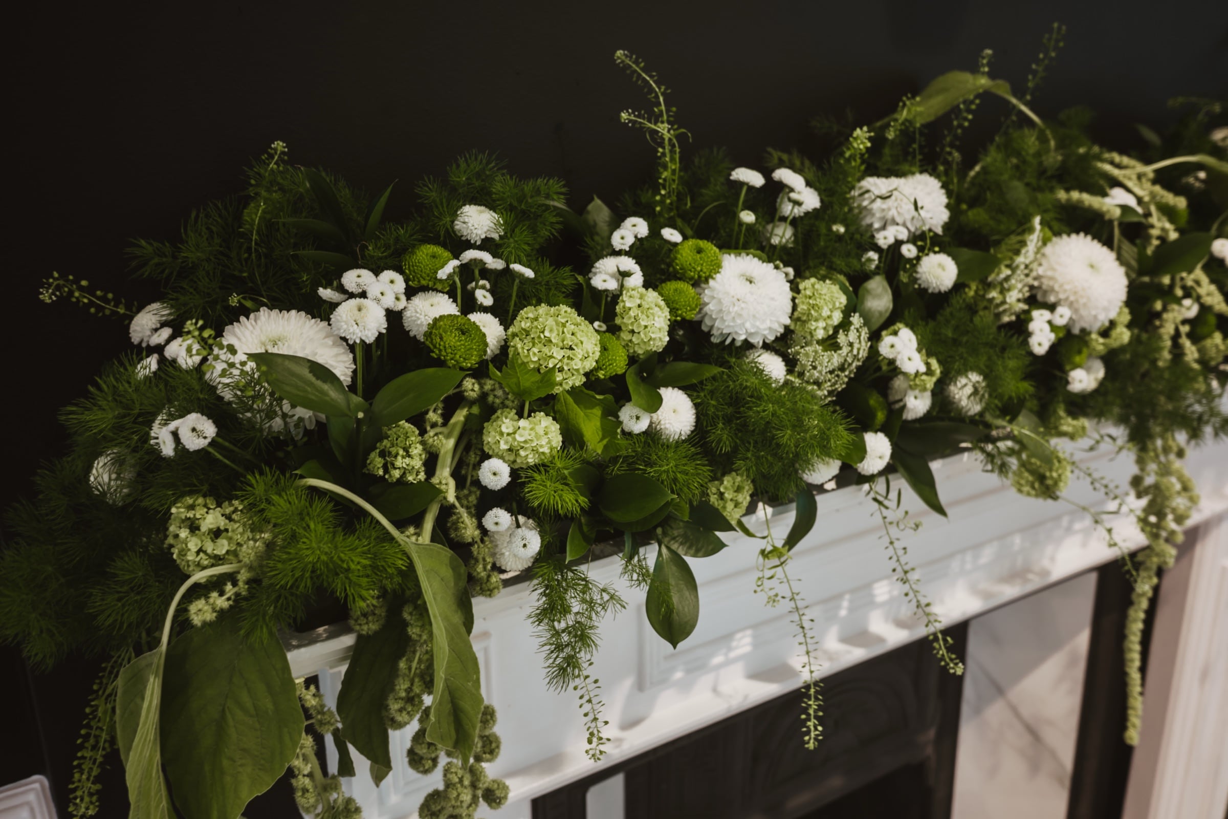 White flowers and greenery running the length of a stone mantel against a dark wall.