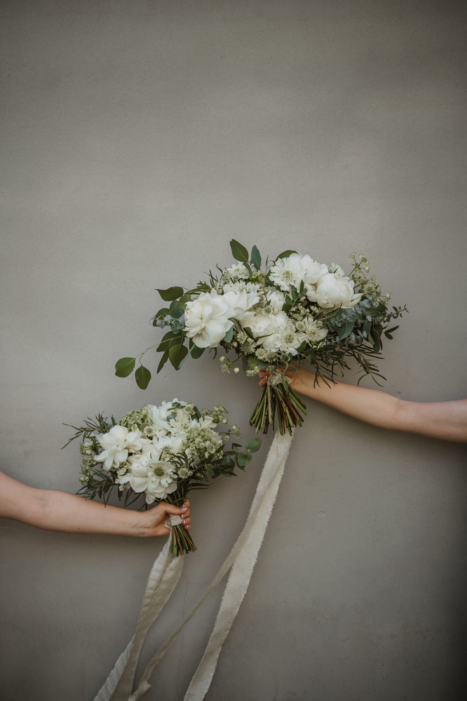 Two hands holding white bouquets against a plain grey wall, ribbons hanging.