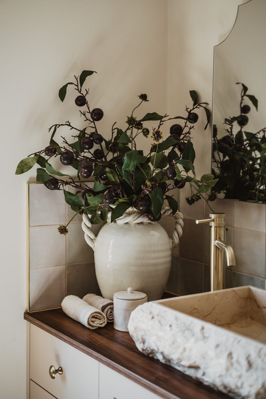 Blackthorn Harvest: plum spray branches with trailing dark berries in a glazed cream urn with twisted handles, on a bathroom vanity beside a stone basin.