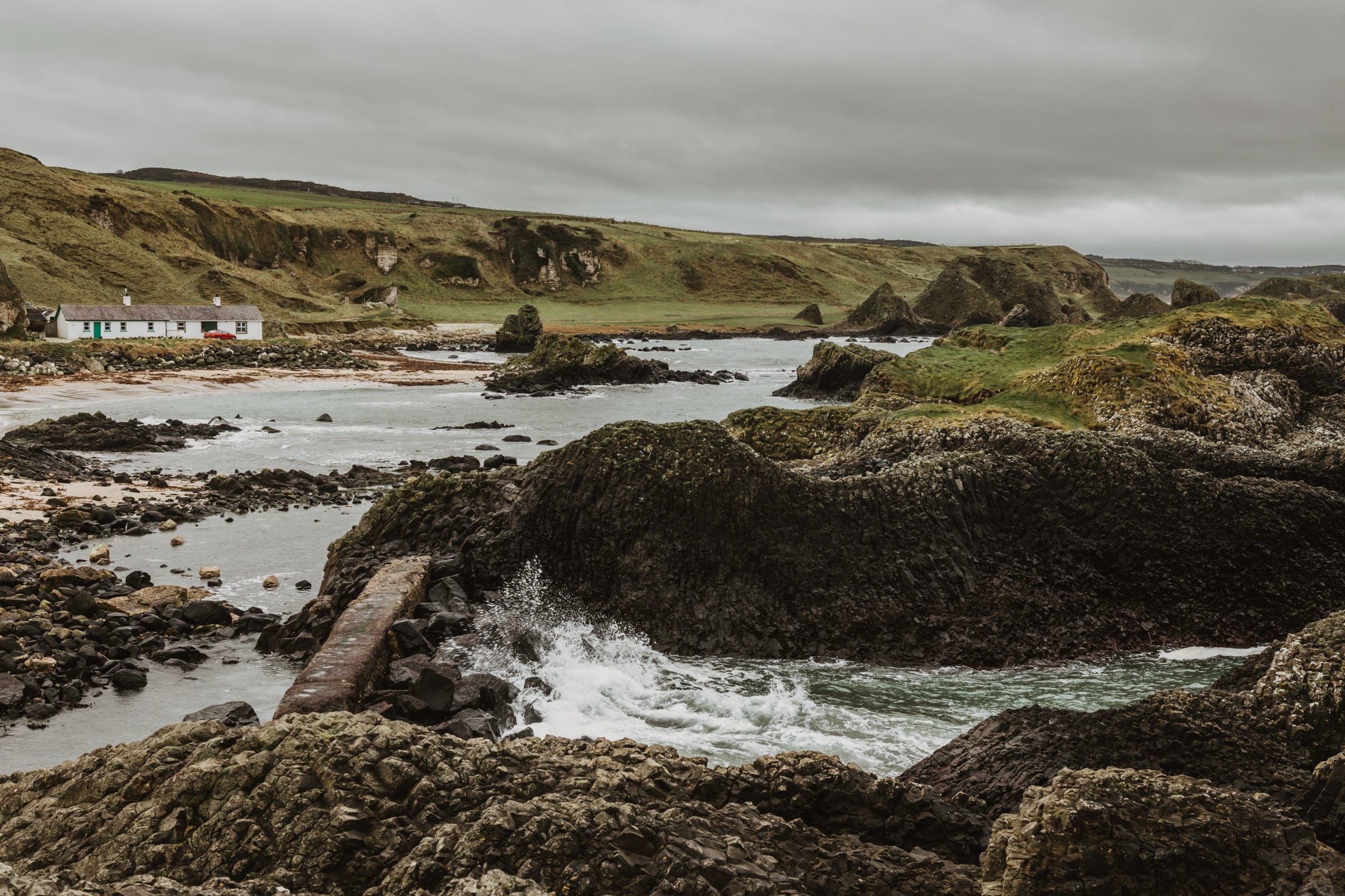 A white cottage above a rocky bay under heavy cloud, with green headland behind and surf breaking over dark basalt in the foreground.