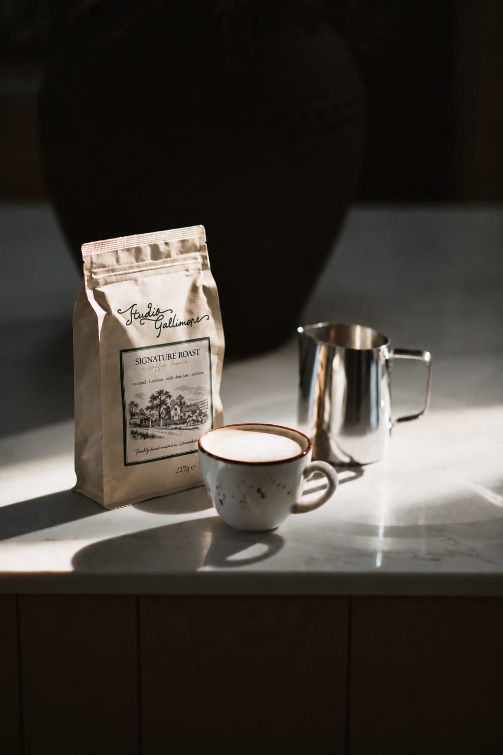A 227 g bag of Studio Gallimore Signature Roast on a marble worktop in low light, with a cappuccino in a speckled cup and a steel milk jug.