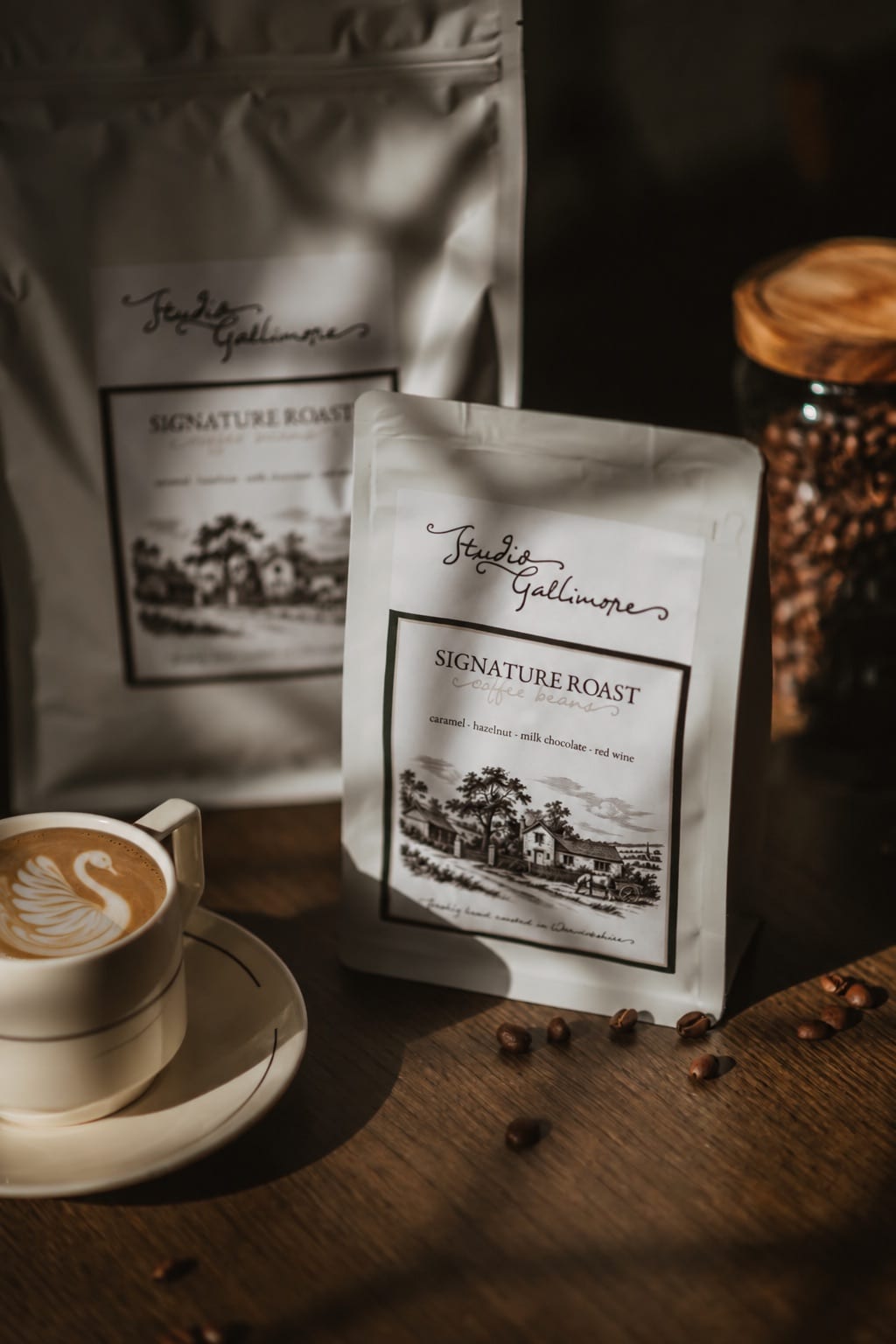 A 227 g bag of Studio Gallimore Signature Roast standing in low light, its label naming caramel, hazelnut, milk chocolate and red wine, with the 1 kg bag behind, a jar of beans and a cup of coffee with a swan in the foam.