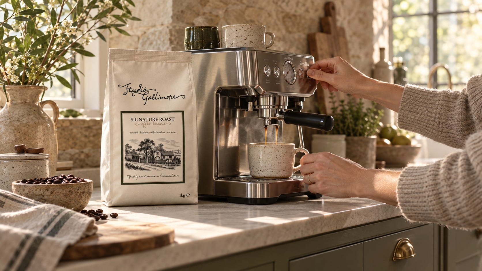 Hands pulling an espresso into a speckled cup at a steel machine on a marble worktop, a 1 kg bag of Studio Gallimore Signature Roast beside it, in morning light.
