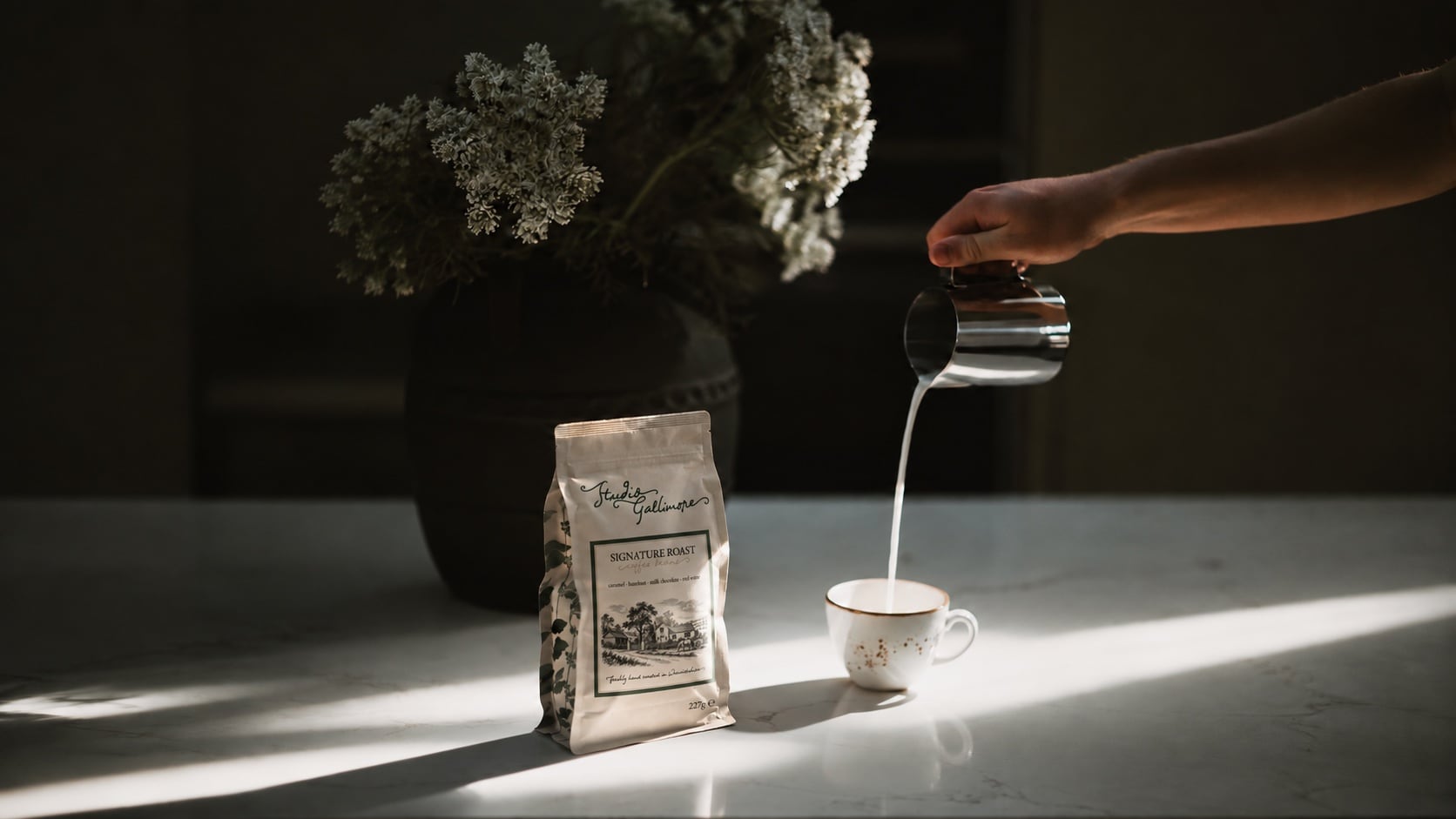 Milk poured from a steel jug into a cup on a marble worktop, a bag of Studio Gallimore Signature Roast standing beside it, white blossom in a dark vase behind.