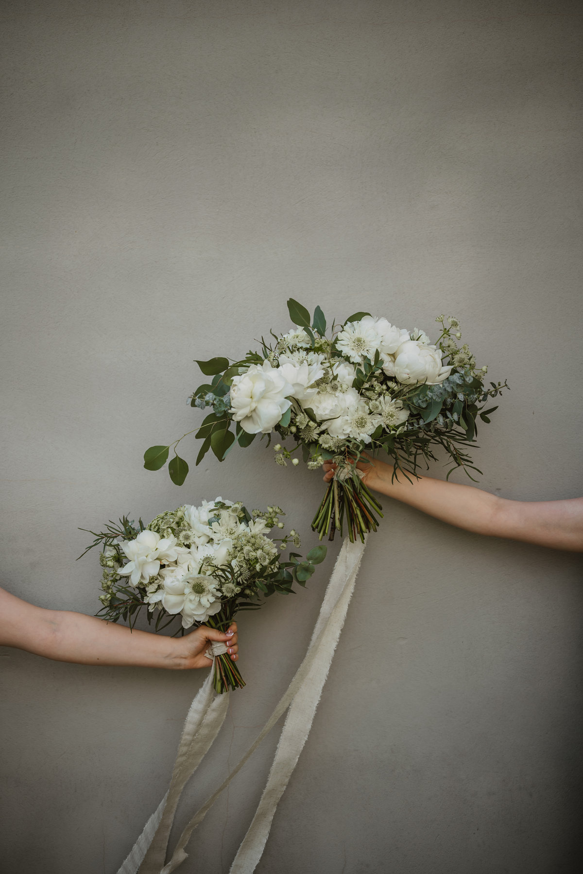 Two hands holding white bouquets against a plain grey wall, ribbons hanging.